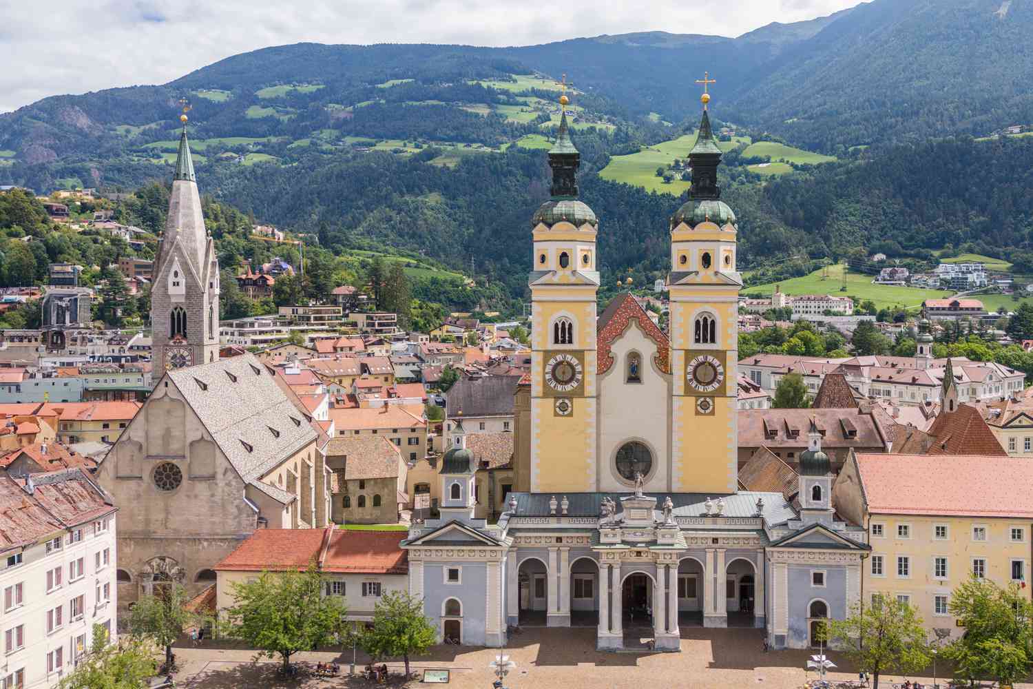 Une vue aérienne montre le centre-ville de Brixen, Italie, y compris une grande église en pierre avec un haut clocher et une église peinte en jaune et bleu avec deux clochers