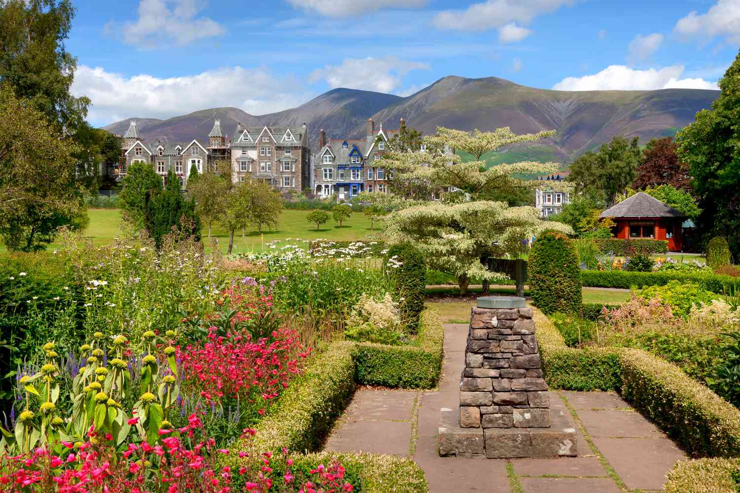 Un jardin traditionnel anglais, avec de grandes fleurs et des statues en pierre, se trouve au premier plan avec une rangée de maisons de ville en arrière-plan et une chaîne de montagnes au loin